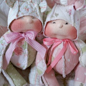 May include: Two handmade fabric dolls dressed in pink and white patchwork clothing with floral patterns. The dolls have pink bows and are sitting on a pink and white quilt.