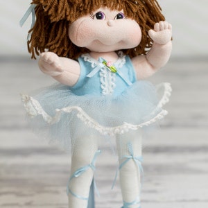 Puede incluir: Una muñeca hecha a mano con pelo de lana marrón, un vestido azul claro con ribete de encaje blanco y zapatos de ballet azul claro. La muñeca está de pie sobre un soporte blanco.