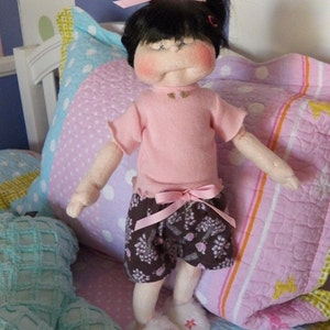 May include: A doll with black hair and pink bow wearing a pink shirt and brown floral shorts. The doll is sitting on a bed with a pink and white patterned quilt.