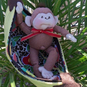 May include: A brown plush monkey with a red bow tie is sitting inside a yellow banana-shaped pouch with a colorful floral pattern.