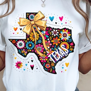 May include: White t-shirt featuring a colorful floral design in the shape of Texas. The design includes a yellow bow, the word "TEXAS", and various flowers and hearts. The shirt is a casual wear item.