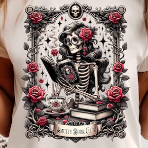 Peut inclure: T-shirt crème avec une illustration détaillée d'un squelette lisant un livre, orné de roses et d'un motif de crâne. Le texte "Smutty Book Club" est affiché sur une bannière. Le design comprend une tasse à thé et des livres.