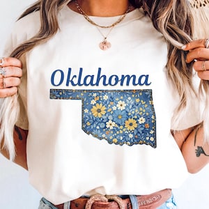 May include: Cream-colored t-shirt featuring the word "Oklahoma" in blue script above a floral-patterned outline of the state. The floral design includes blue, yellow, and white flowers. The shirt is paired with blue jeans and a brown belt.