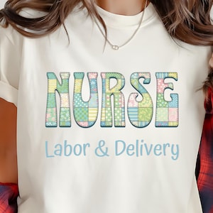 Puede incluir: Camiseta color crema con la palabra "NURSE" en letras grandes y coloridas, estilo patchwork. Debajo, las palabras "Labor & Delivery" están escritas en fuente azul claro. La camiseta tiene mangas cortas.