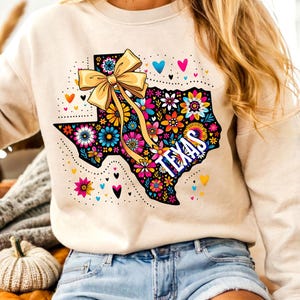 Puede incluir: Sudadera beige con un dise&ntilde;o floral colorido del estado de Texas, un lazo amarillo y la palabra "TEXAS". El dise&ntilde;o incluye corazones y detalles punteados. La sudadera se combina con pantalones cortos de mezclilla.