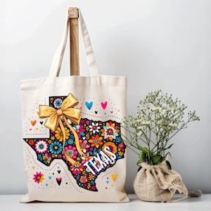 Puede incluir: Un bolso tote de color crema con un dise&ntilde;o floral que representa el estado de Texas. El dise&ntilde;o incluye un lazo amarillo, corazones y la palabra "TEXAS" en blanco. El bolso tiene un asa de madera.