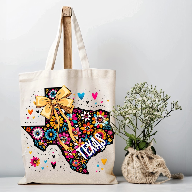Puede incluir: Un bolso tote de color crema con un dise&ntilde;o floral que representa el estado de Texas. El dise&ntilde;o incluye un lazo amarillo, corazones y la palabra "TEXAS" en blanco. El bolso tiene un asa de madera.