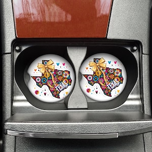 Puede incluir: Dos posavasos blancos para coche con un dise&ntilde;o colorido de Texas. El dise&ntilde;o presenta un contorno floral de Texas con un lazo dorado y la palabra "Texas". Los posavasos est&aacute;n en los portavasos de un coche.