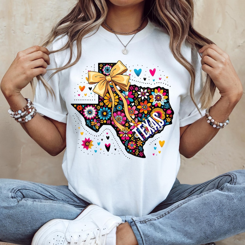 May include: White t-shirt featuring a colorful floral design in the shape of Texas. The design includes a yellow bow, the word "TEXAS", and various flowers and hearts. The shirt is a casual wear item.