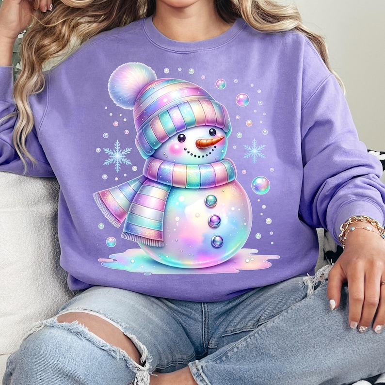 May include: A lilac-colored sweatshirt featuring a whimsical snowman illustration. The snowman has a rainbow-colored hat, scarf, and buttons. Snowflakes and bubbles surround the snowman, creating a festive and colorful design.