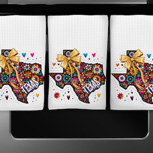 Puede incluir: Tres pa&ntilde;os de cocina blancos con un dise&ntilde;o en forma de Texas. El dise&ntilde;o presenta un colorido estampado floral, la palabra "TEXAS" y un lazo dorado. Corazones y flores de varios colores rodean el contorno de Texas.