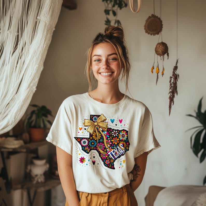 Puede incluir: Camiseta color crema con un dise&ntilde;o floral colorido del contorno del estado de Texas, con un lazo dorado y la palabra "TEXAS". El dise&ntilde;o incluye corazones y flores de varios colores.