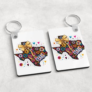 Puede incluir: Dos llaveros rectangulares blancos con un dise&ntilde;o floral del contorno del estado de Texas y un lazo dorado. La palabra "TEXAS" est&aacute; impresa en el dise&ntilde;o. Cada llavero tiene un anillo plateado.