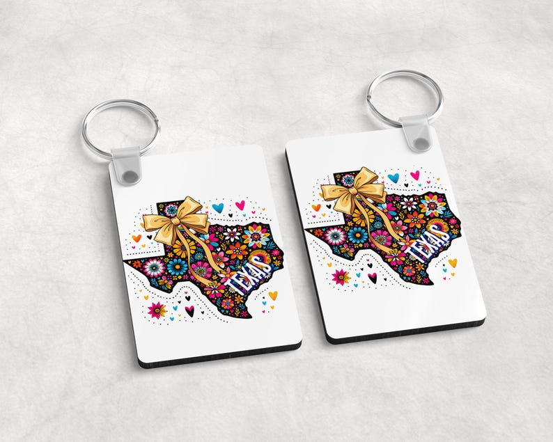Puede incluir: Dos llaveros rectangulares blancos con un dise&ntilde;o floral del contorno del estado de Texas y un lazo dorado. La palabra "TEXAS" est&aacute; impresa en el dise&ntilde;o. Cada llavero tiene un anillo plateado.