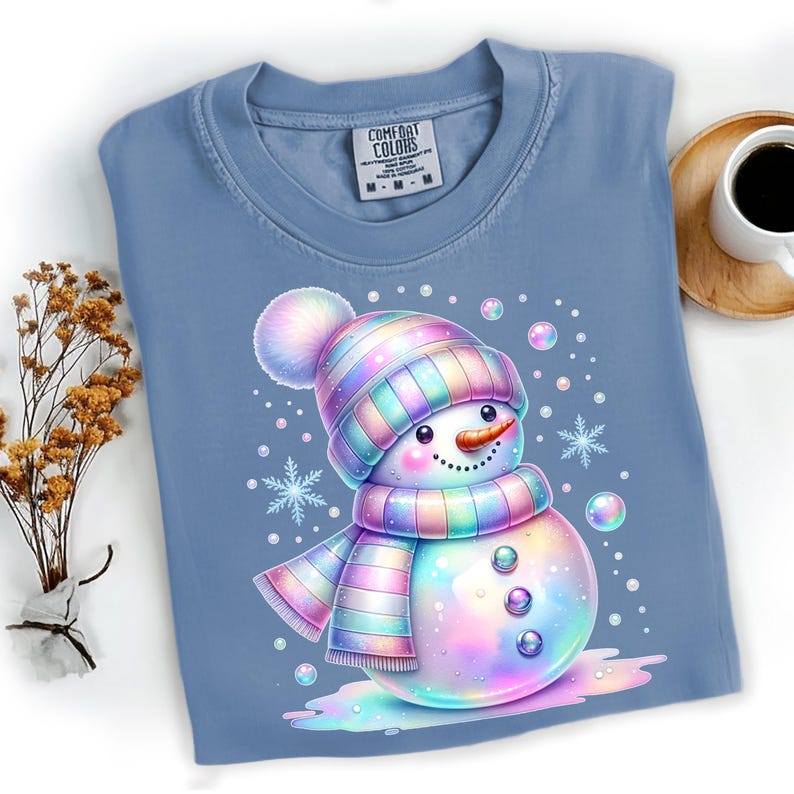 Puede incluir: Camiseta azul Comfort Colors con un dise&ntilde;o de mu&ntilde;eco de nieve colorido. El mu&ntilde;eco de nieve lleva un gorro y una bufanda a rayas, una nariz de zanahoria y ojos de bot&oacute;n. Copos de nieve y burbujas rodean al mu&ntilde;eco de nieve.