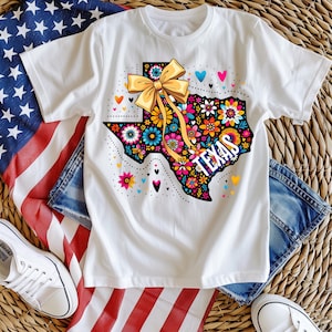 Puede incluir: Camiseta blanca con un dise&ntilde;o floral colorido de Texas, un lazo amarillo y la palabra "TEXAS". El dise&ntilde;o incluye varias flores, corazones y un contorno punteado del estado. La camiseta se combina con pantalones cortos de mezclilla y zapatillas blancas.