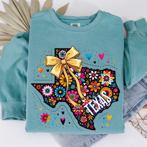Puede incluir: Una sudadera de color verde azulado con un dise&ntilde;o floral colorido del contorno de Texas y un gran lazo amarillo. La palabra "TEXAS" est&aacute; impresa en blanco. Peque&ntilde;os corazones de varios colores est&aacute;n dispersos alrededor del dise&ntilde;o.