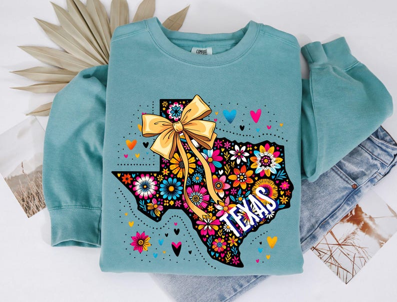 Puede incluir: Una sudadera de color verde azulado con un dise&ntilde;o floral colorido del contorno de Texas y un gran lazo amarillo. La palabra "TEXAS" est&aacute; impresa en blanco. Peque&ntilde;os corazones de varios colores est&aacute;n dispersos alrededor del dise&ntilde;o.