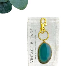 May include: A teal agate stone keychain with a gold metal ring and clasp. The keychain is packaged in a small white cloth bag with the text "Vintage Blonde" and a website address.