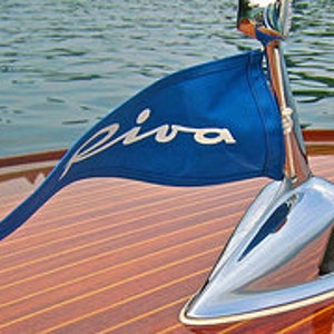 Puede incluir: Una bandera azul con la palabra "Riva" en letras blancas, unida a un asta de bandera cromada en un barco de madera.