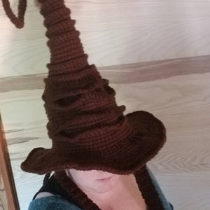 Peut inclure: Un chapeau de sorcier marron crocheté avec une couronne haute et pointue et un large bord. Le chapeau a un aspect texturé et de longues attaches tressées. Le chapeau est de couleur marron foncé.