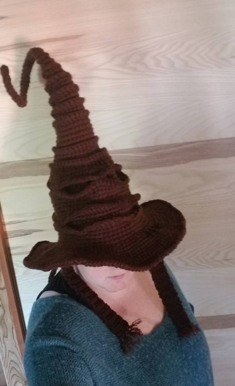 May include: A brown, crocheted wizard hat with a tall, pointed crown and a wide brim. The hat has a textured appearance and long, braided ties. The hat is a dark brown color.