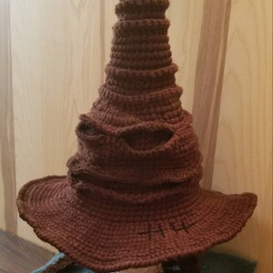 May include: A brown, crocheted Sorting Hat with a wide brim and a curved, horn-like detail. The hat features a textured surface and is designed to resemble the iconic hat from the wizarding world. The hat is a handmade item.