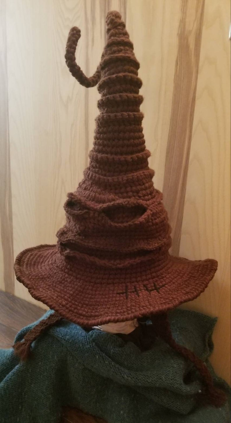 May include: A brown, crocheted Sorting Hat with a wide brim and a curved, horn-like detail. The hat features a textured surface and is designed to resemble the iconic hat from the wizarding world. The hat is a handmade item.