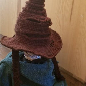 May include: A handmade, brown crochet Sorting Hat with a wide brim and a tall, pointed crown. The hat has a textured appearance and is resting on a blue-green fabric. The hat is a replica from the Harry Potter series.