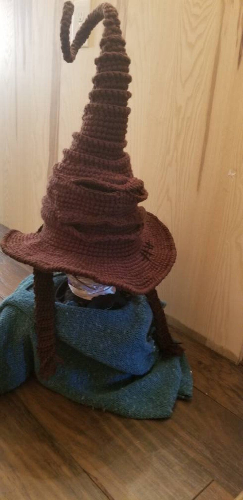 May include: A handmade, brown crochet Sorting Hat with a wide brim and a tall, pointed crown. The hat has a textured appearance and is resting on a blue-green fabric. The hat is a replica from the Harry Potter series.