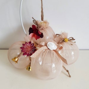May include: Three beige tulle Christmas baubles with gold bells and floral embellishments. The baubles are decorated with white polka dots, red and white flowers, and a white bow with a white pearl centre.