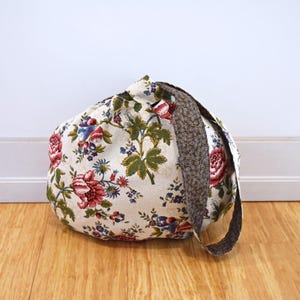 May include: A white fabric tote bag with a floral print featuring red, blue, and green flowers. The bag has a brown fabric strap.