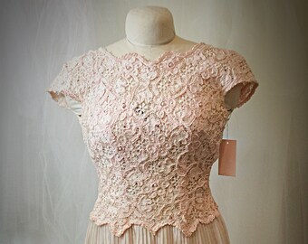 May include: A vintage blush-colored dress displayed on a mannequin. The bodice features intricate lace detailing, short sleeves, and a scalloped neckline. The skirt is pleated, creating a flowing silhouette. A tag hangs from the dress.