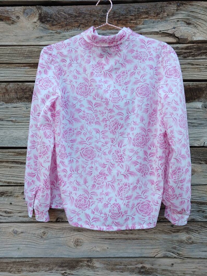 May include: A white blouse with a pink floral pattern. The blouse has a high neck and long sleeves.