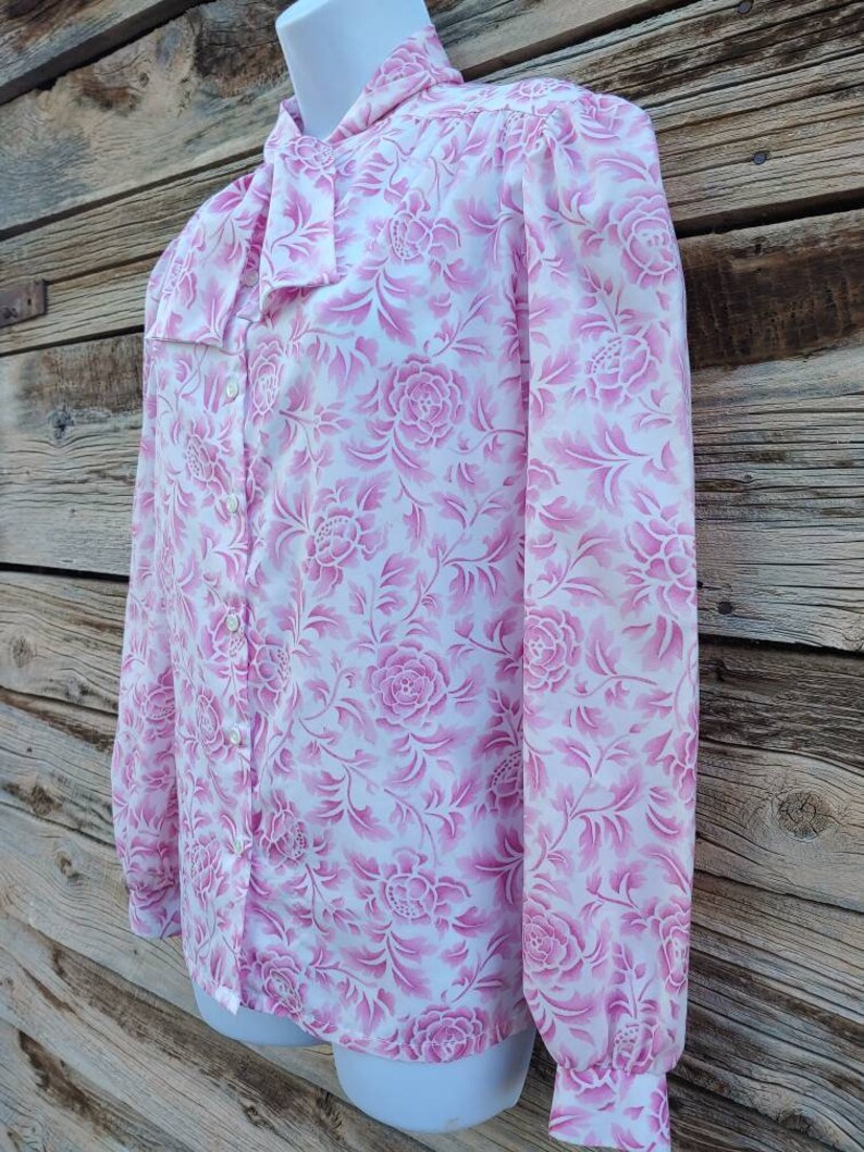 May include: A white long-sleeve blouse with a pink floral pattern. The blouse has a bow tie collar and button closure.