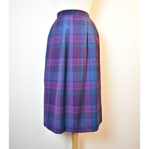 May include: A knee-length skirt in a plaid pattern, featuring shades of purple, blue, and pink. The skirt is displayed on a mannequin, showcasing its pleated design and tailored fit. The fabric appears to be a medium-weight material.