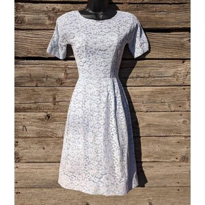 May include: A light blue knee-length dress featuring a delicate floral lace overlay, short sleeves, and a round neckline. The garment has a fitted bodice that transitions into a slightly flared skirt, showcasing a consistent lace pattern throughout.