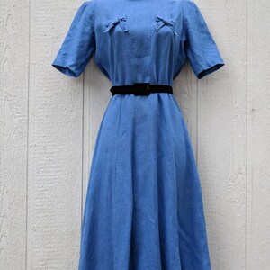 May include: A light blue vintage dress with a fitted bodice and a full skirt. The dress has a round neckline and short sleeves. It features two bows on the bodice and a black belt.