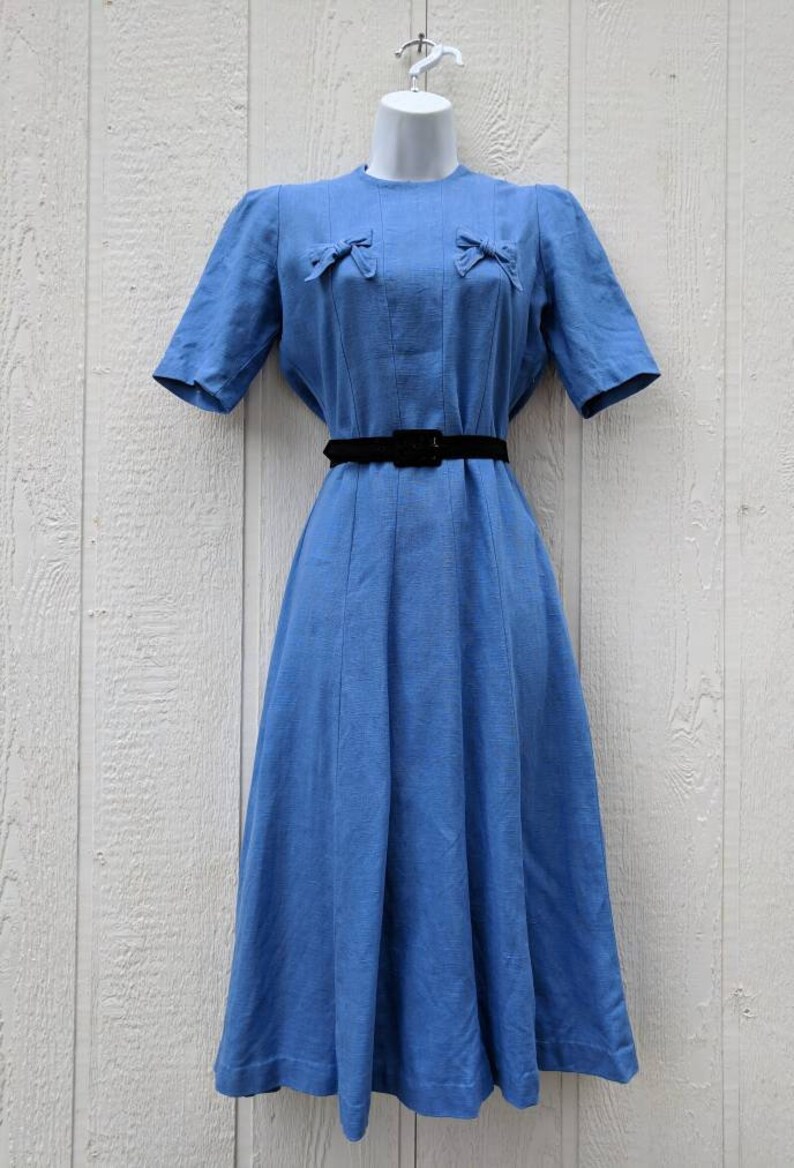 May include: A light blue vintage dress with a fitted bodice and a full skirt. The dress has a round neckline and short sleeves. It features two bows on the bodice and a black belt.