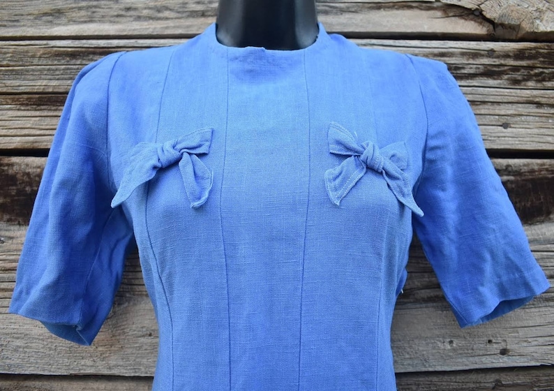May include: A blue linen dress with a round neckline and short sleeves. The dress has two bows on the front, one on each side of the neckline.