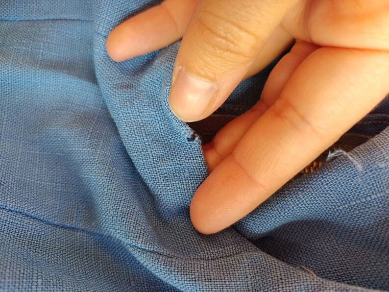 May include: Close-up of a blue fabric with a small tear in the material. A finger is shown pointing at the tear.