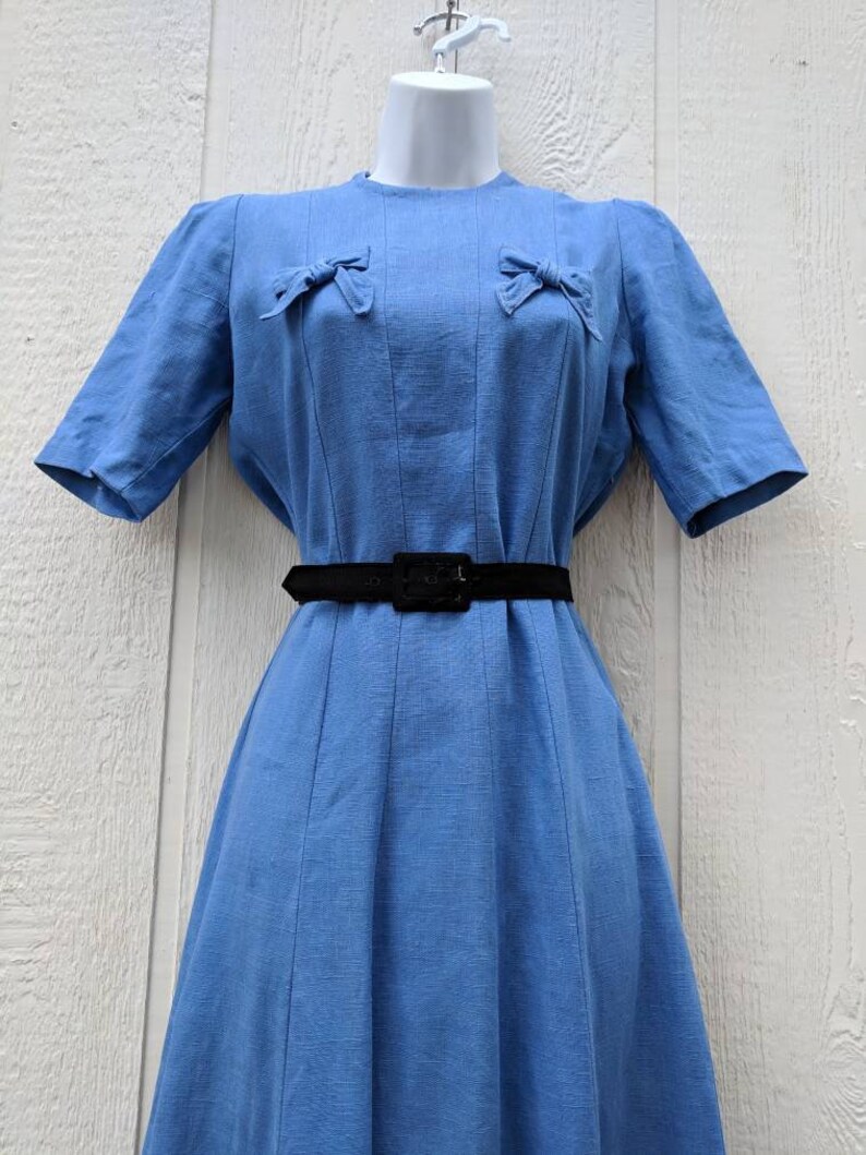 May include: A light blue dress with a black belt and two bows on the chest. The dress has a fitted bodice and a full skirt.