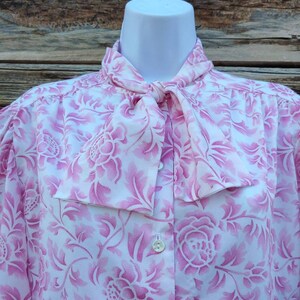 May include: A white blouse with a pink floral pattern and a bow tie. The blouse has a button-up front and long sleeves.