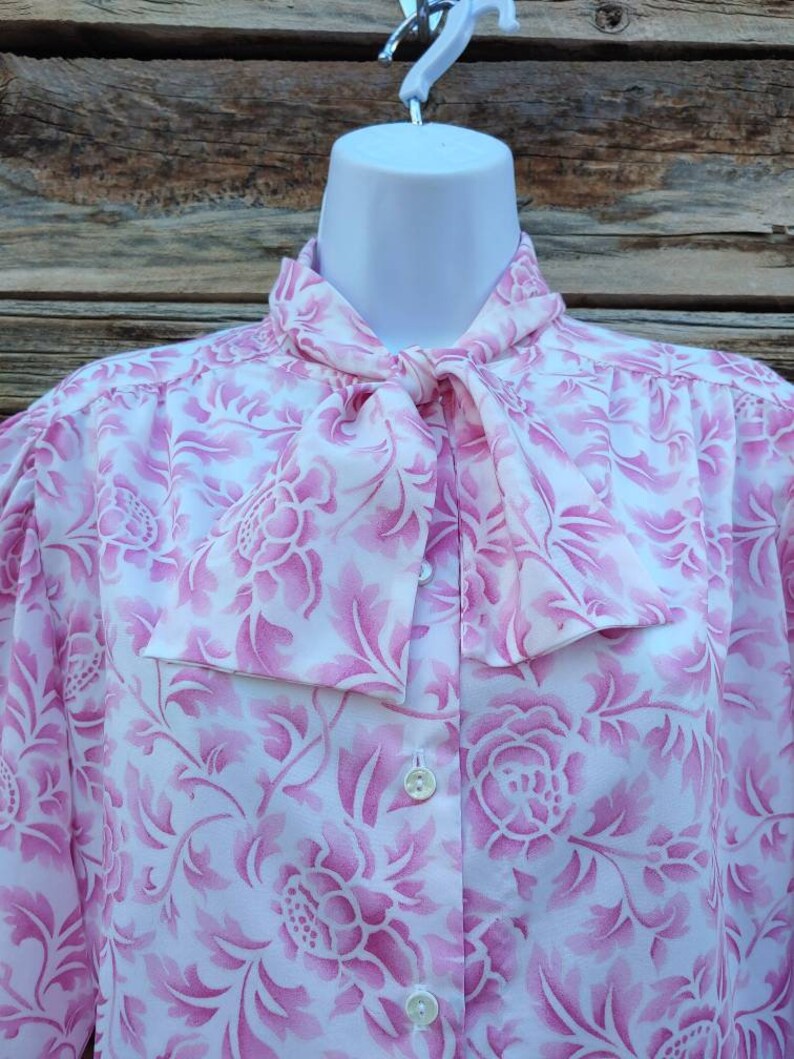 May include: A white blouse with a pink floral pattern and a bow tie. The blouse has a button-up front and long sleeves.