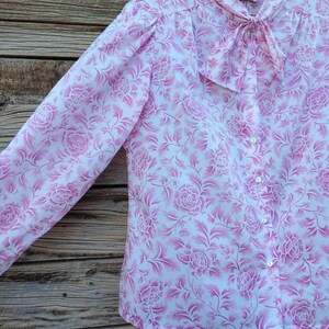 May include: A white blouse with a pink floral pattern and a bow tie at the collar. The blouse has a button-up front and long sleeves.