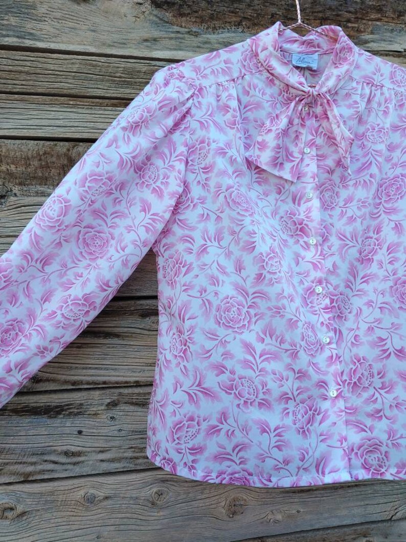 May include: A white blouse with a pink floral pattern and a bow tie at the collar. The blouse has a button-up front and long sleeves.