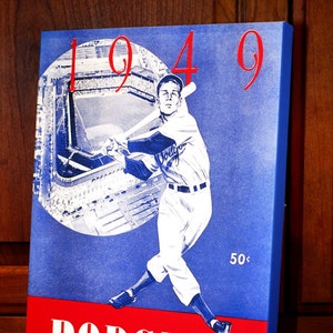 May include: A vintage-style canvas print featuring a baseball player in a Dodgers uniform, poised to swing. The image is set against a blue background with the year "1949" in red. The word "DODGERS" is displayed in white on a red banner.