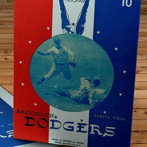 Könnte beinhalten: Eine Vintage-Scorecard der Brooklyn Dodgers aus dem Jahr 1943 in den Farben Blau, Weiß und Rot. Die Karte zeigt ein Foto eines laufenden Baseballspiels mit dem Text "OFFICIAL SCORE CARD 1943 PROGRAM 10c" oben und "BROOKLYN DODGERS EBBETS FIELD" unten.