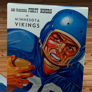 Può includere: Una copertina di programma di football americano vintage con un primo piano di un giocatore di football in una maglia blu e un casco, che tiene un pallone da football. Il testo sulla copertina recita "SAN FRANCISCO FORTY NINERS vs. MINNESOTA VIKINGS" e "NOVEMBER 26, 1961".