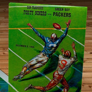 Może przedstawiać: Okładka programu meczu futbolu amerykańskiego w stylu vintage, w kolorze zielonym i białym, przedstawiająca San Francisco Forty Niners przeciwko Green Bay Packers. Okładka programu przedstawia dwóch graczy futbolu amerykańskiego w akcji, jeden w niebieskim stroju, a drugi w brązowym. Tekst na okładce brzmi "San Francisco Forty Niners vs. Green Bay Packers" i "8 grudnia 1956".
